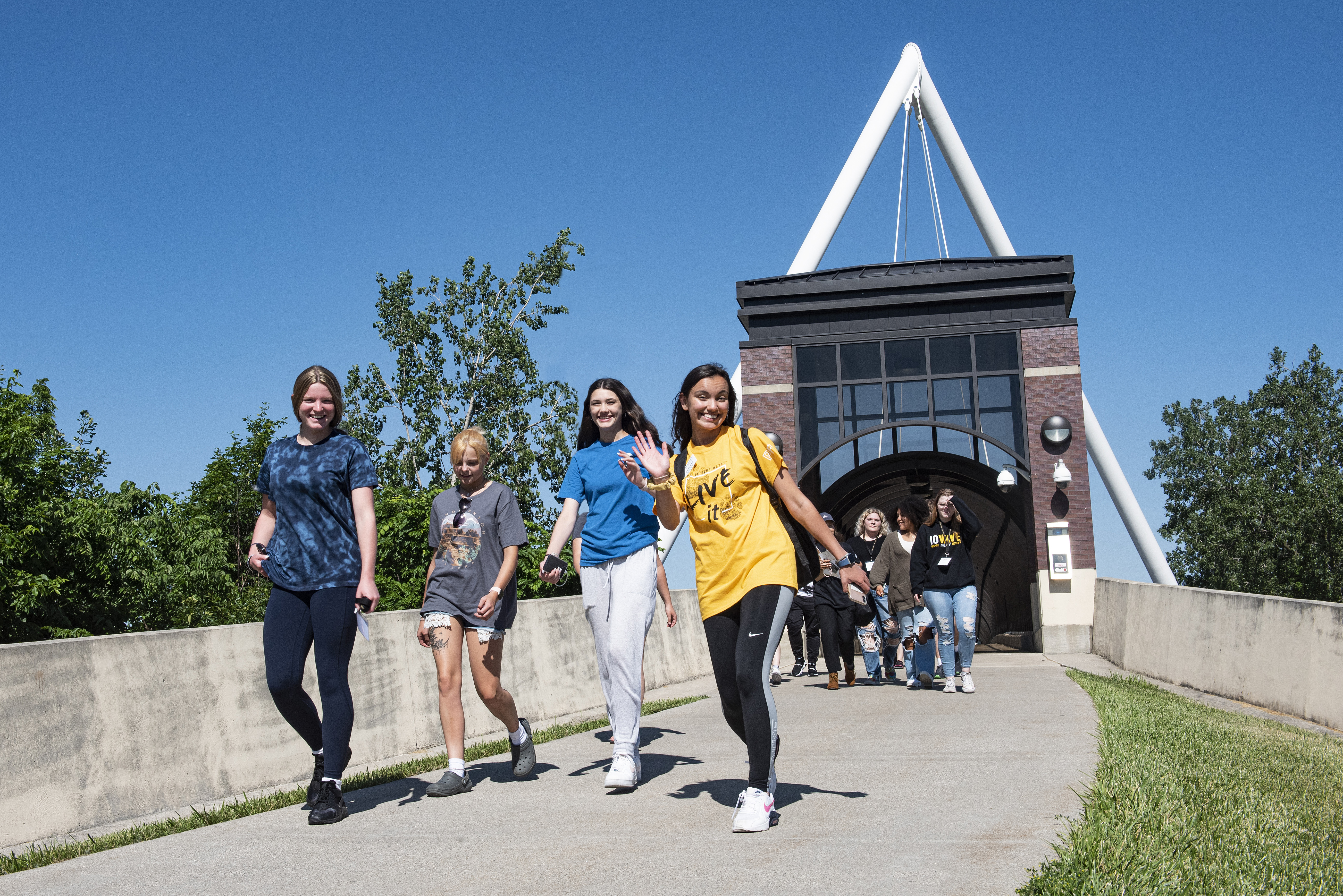 A group of smiling students walk on a sunny day across a pathway toward the camera. They are in front of a brick building with a large metal arch.